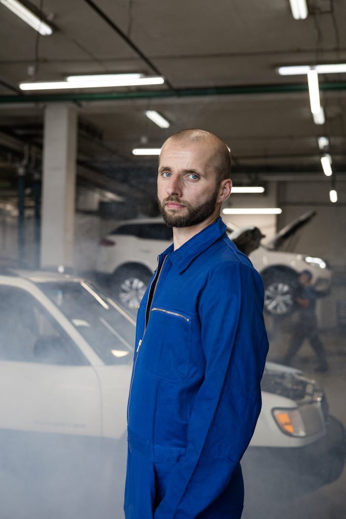 Mechanic wearing a blue coverall in an auto repair shop with cars in the background.